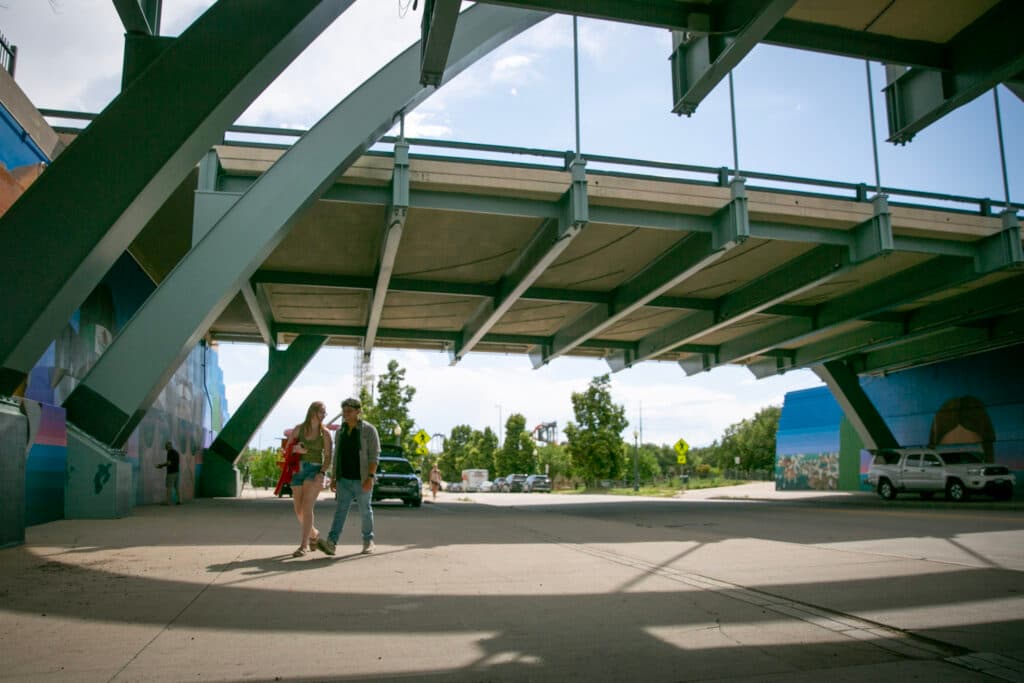 A young man and woman stroll down a broad sidewalk, road to their left, murals on either side of the road. Emanuel Martinez is in the shadows in the background, painting the wall.