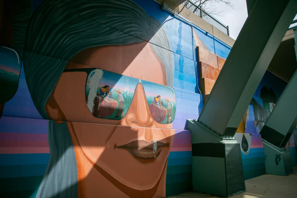 A sunglassed figure with dark skin and long black hair from Emanuel Martinez's "Confluent People." The person's lenses reflect a traditional Mexican ballet folklorico with a man and a woman standing in a field, preparing to dance.