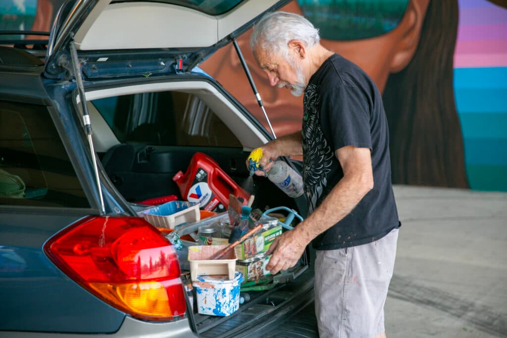 Emanuel Martinez uses a spray bottle to mist his paint brush and tray, set up in the back of a Subaru hatchback. Behind him you can see one of the faces from the most iconic panel of his "Confluent People" mural.