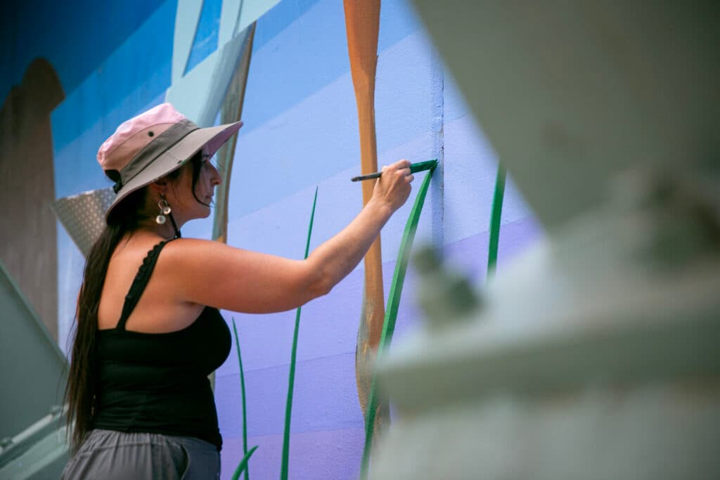 Bianca Maestas paints the tip of a large, green blade of grass. She's wearing a pink bucket hat and a black tank top.