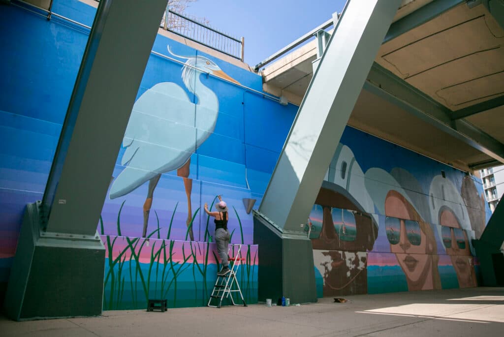 Bianca Maestas stands on a ladder and paints the tip of a large, green blade of grass. A large, painted great blue heron looms above her and three painted heads look at her from the wall on her right.