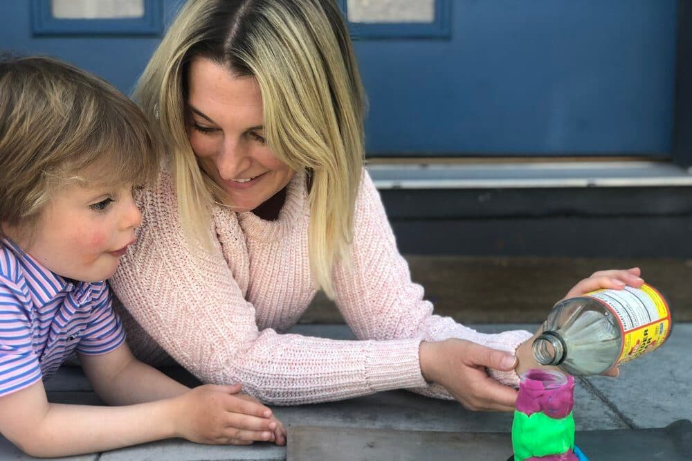 A blonde white woman lays on the ground with a child. She is poised to pour apple cider vinegar into a colorful volcano science experiment.