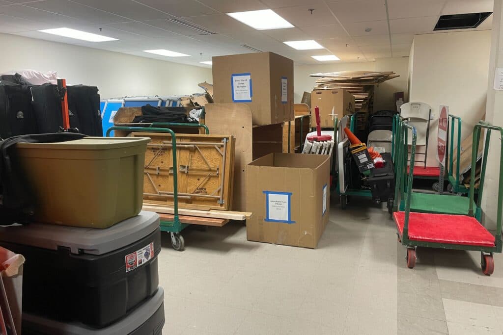 A room lit by bright overhead fluorescent lights is packed with plastic and cardboard storage boxes.