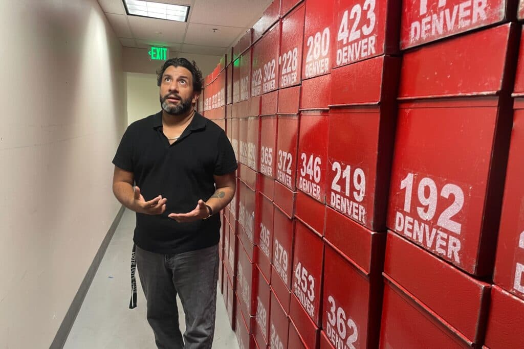 A Latino man wearing a black polo and grey shirts walks beside a hallway packed with rows of red boxes.