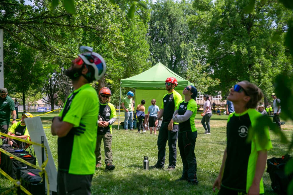 A crowd of competitors in vibrant yellow/green shirts look up at a tree, watching Wyatt Chipps (out of frame) competing in the throwline event.