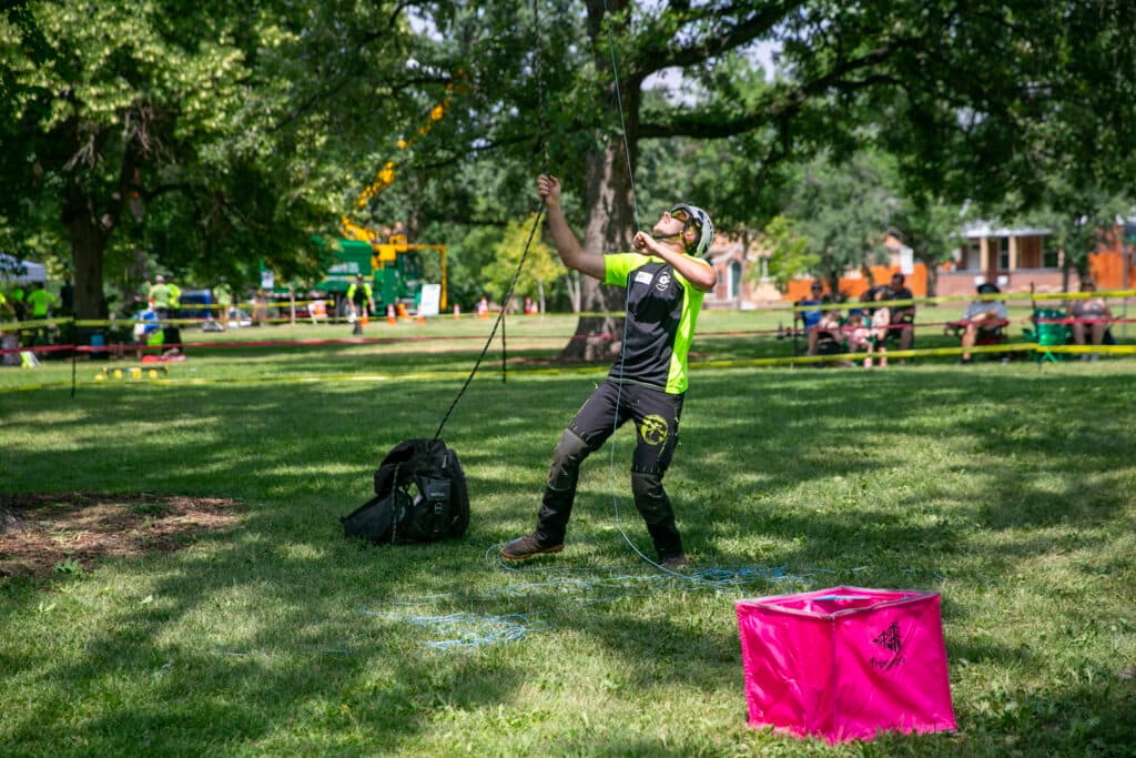 Wyatt Chipps looks up at the canopy of a tree as each hand grasps a blue rope that he has thrown up and out of view.