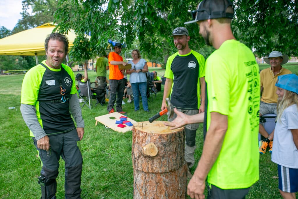 Patrick O'Meara stands in a circle around a stump with nails in it. He's playing hammerschlagen with two other men, one of whom is actively flipping the hammer over backwards before trying to hit one of the nails in the stump.