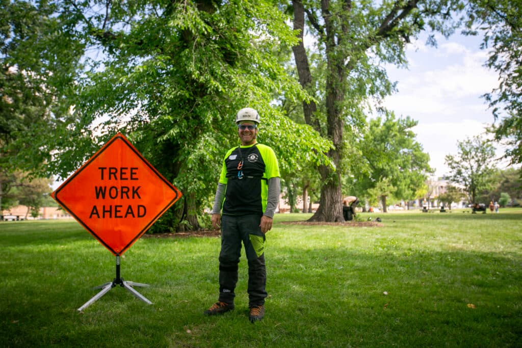 Patrck O'Meara wears a white helmet, vibrant shirt, and safety googles while posing next to an orange "TREE WORK AHEAD" sign in a park.