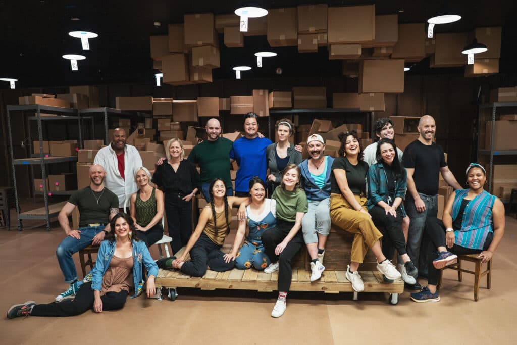 A group of 17 performers pose together on the set of Sweet & Lucky: Echo. A room of cardboard boxes is visible behind them.