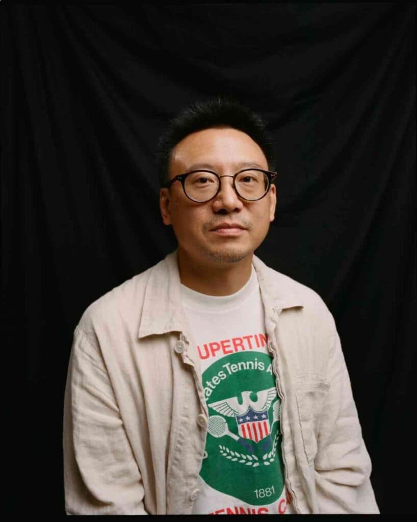 A man sits in front of a black screen with glasses on staring at the camera unsmiling