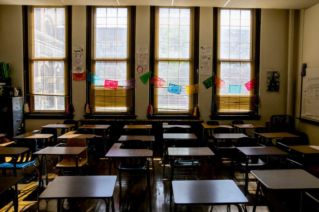 An empty classroom with multiple desks and chairs arranged in rows. The room is lined with windows, providing natural light.