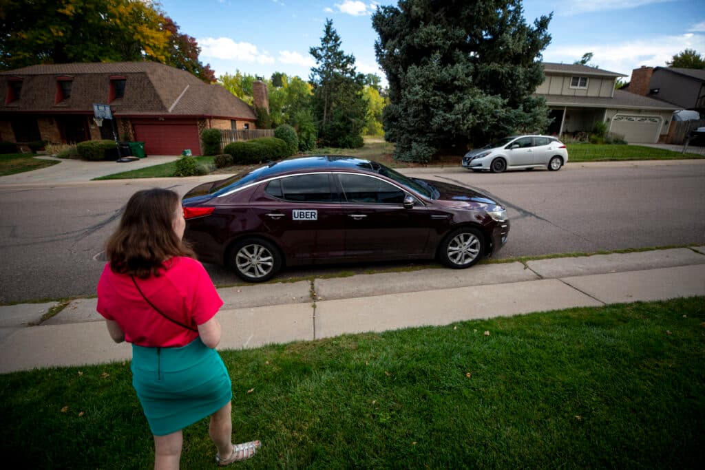 A woman in a red shirt and a teal skirt walks across grass towards a dark car parked in the street. A sign reads UBER along its side.
