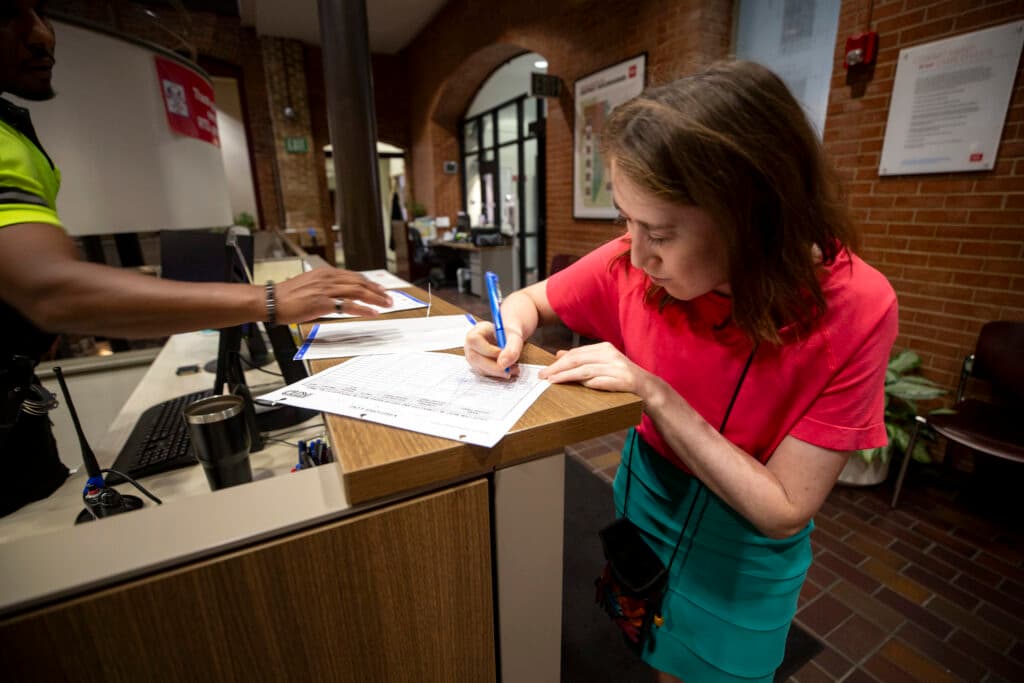 A woman in a red shirt and a teal skirt writes on a sheet of lined paper on a front office desk. A man in a reflective shirt reaches in from the left side of the frame.