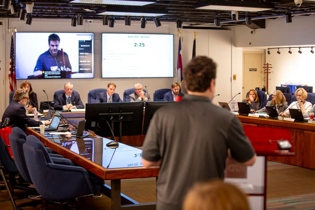 A crowd of people in suit jackets sit around a series of tables, looking seriously. Before them is a man, out of focus, facing them from a podium in the foreground. Behind the stern faces, to one side, is a giant TV that reveals the speaker's face, a man holding a red-handled walking stick who, also looking serious, speaks into a microphone.