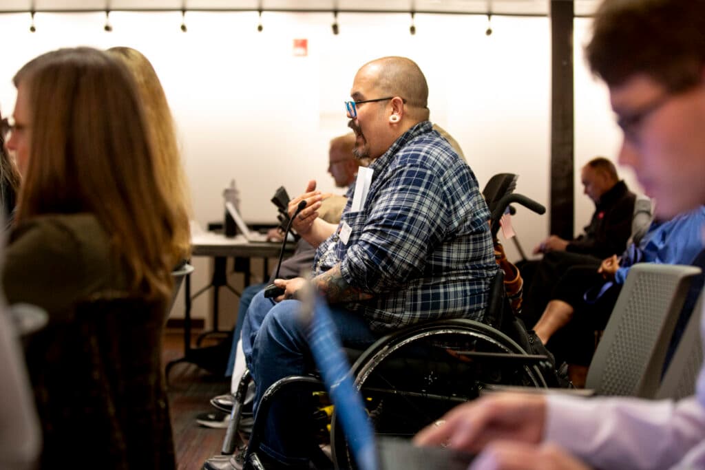A man in a plaid shirt speaks with his hands, possibly shouting, as he holds a microphone and addresses an audience out of frame. He's seated on a wheelchair, surrounded by people looking in the same direction or typing away on laptops.