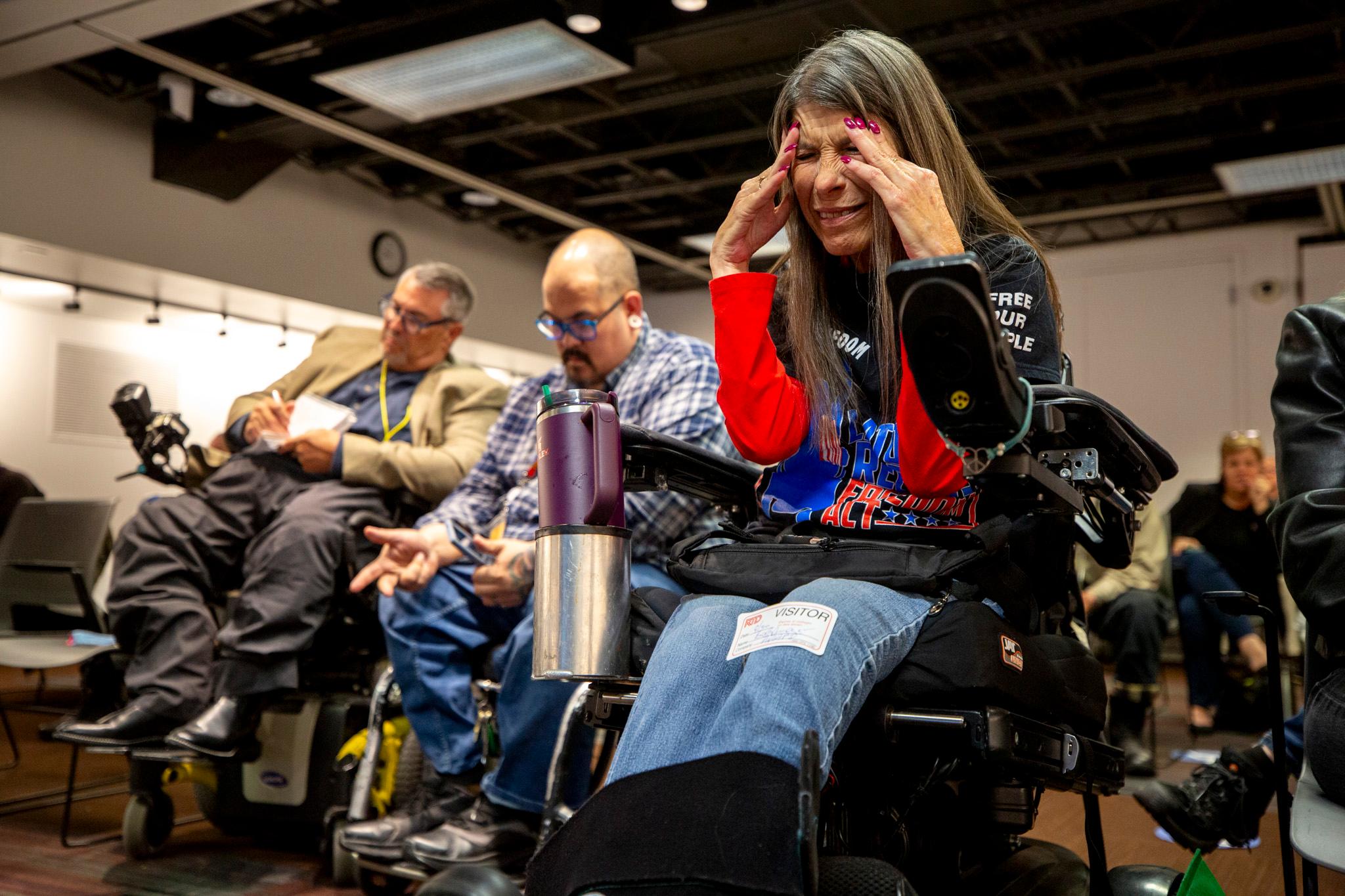 A woman with bright red nails grips her skull with an anguished look on her face. She sits in a room surrounded by people; some, like her, sit in wheelchairs.