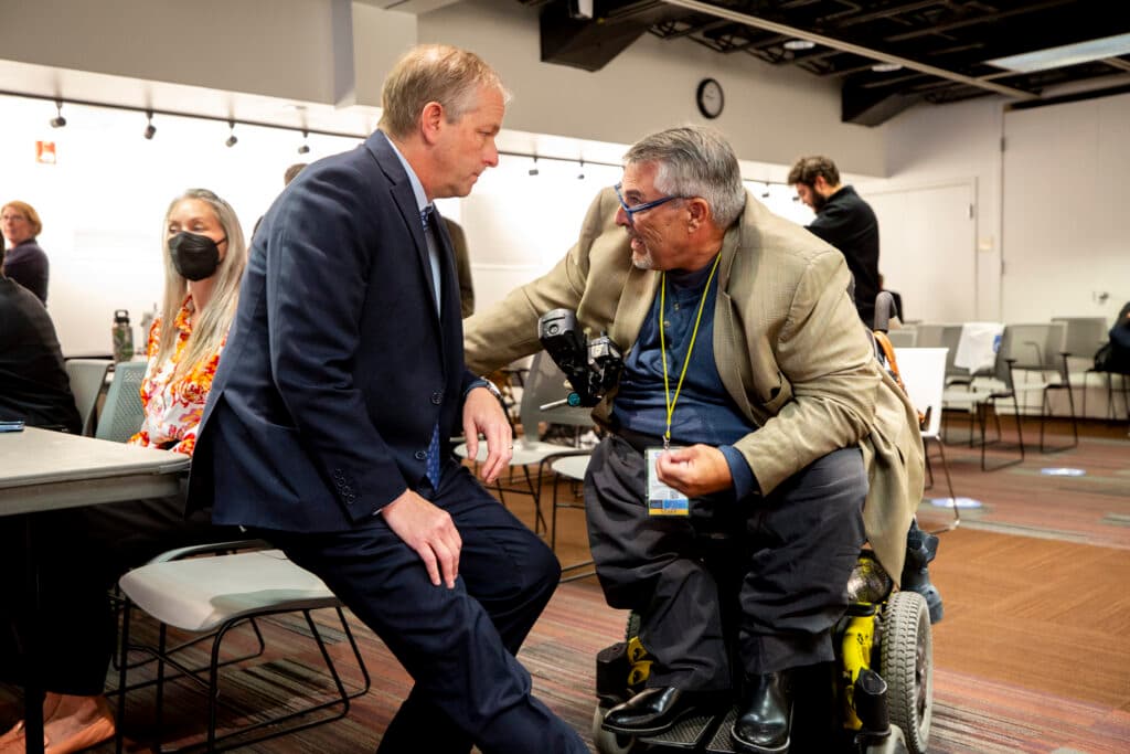A man in a beige jacket leans out of a wheelchair to grab another man, wearing a blue suit, as he speaks with apparent passion.