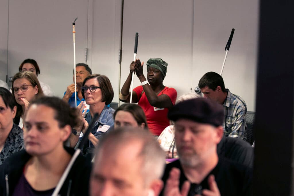 A woman in a red shirt and a green head wrapping holds onto a black-handled walking stick as she brings her other hand up to clap.