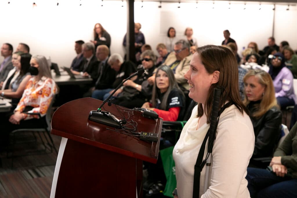 A woman in a white shirt stands at a podium, speaking into a microphone as people behind her watch as she gives public comment.