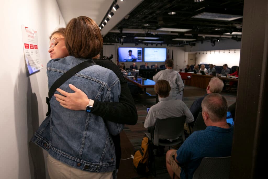 A woman in glasses looks solemn as she embraces another woman, her face peering over the other's shoulder. Behind them is a room full of people sitting around an official-looking table.