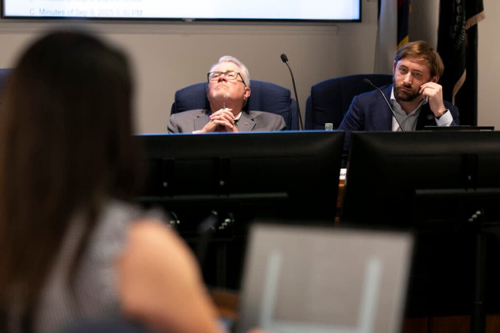 Two men in suits sit behind screens as someone stands before them, way out of focus in the foreground, apparently speaking. One clasps his hands together, leaning back, and stares into the distance. The other stares at her, leaning on his propped arm.