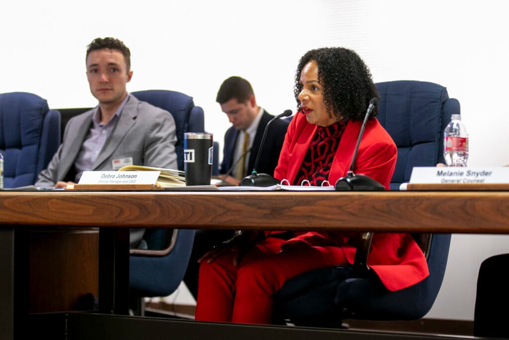 A woman in a cherry red suit slumps to speak into a mic before her, seated behind a mahogany table.