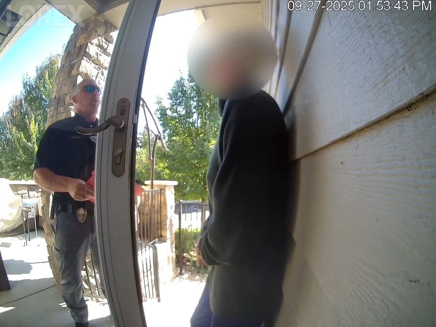 A man in a police uniform is standing and talking to a person, whose face is blurred, right outside their front doorway.