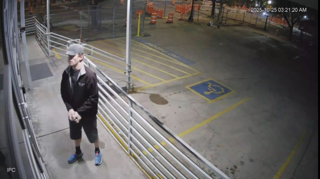 A man is standing outside a business at night, wearing a black jacket, black shorts and a grey baseball cap.The parking lot is empty, and there are no other people visible in the scene.