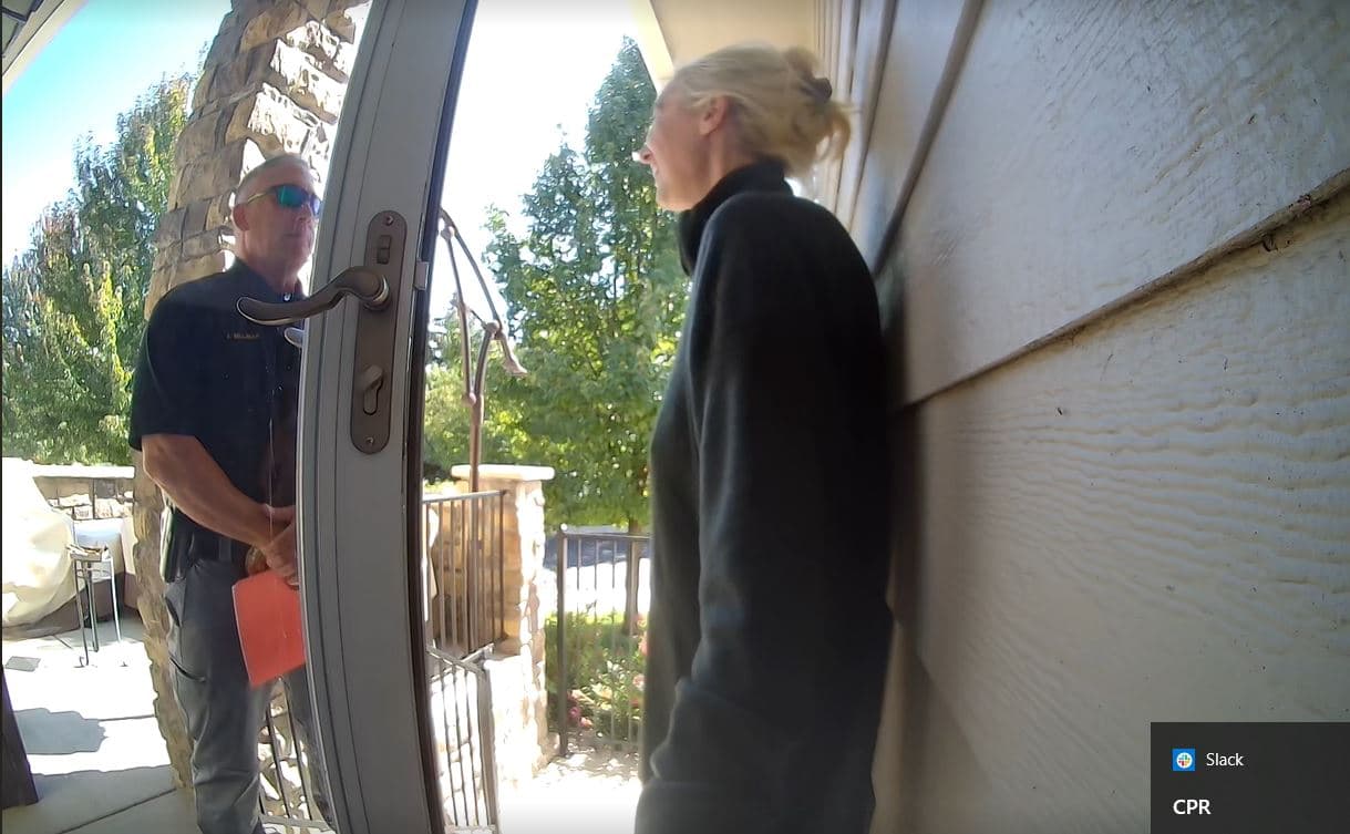 A man in a police uniform is standing and talking to a blond woman on their front porch. A glass-paneled front door is open in between them.