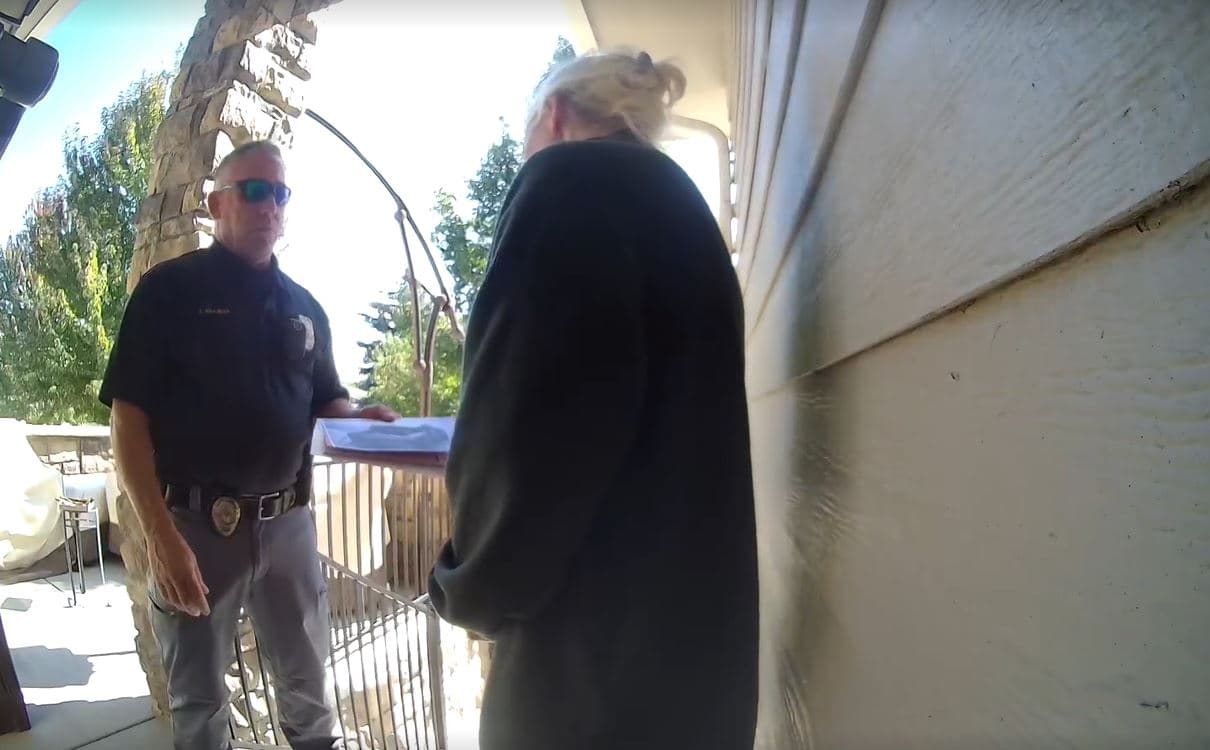 A man in a police uniform is standing and talking to a blond woman on their front porch. A glass-paneled front door is open in between them.