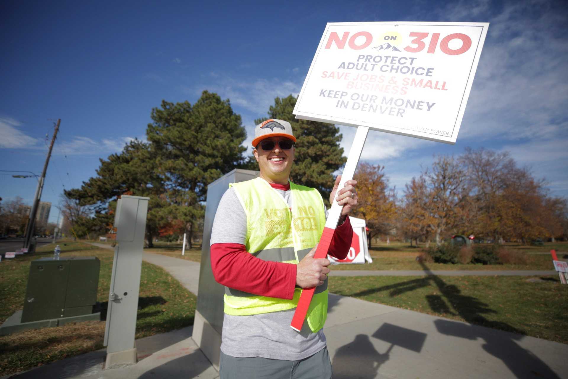 A man, smiling and wearing a yellow safety vest, holds a &quot;No on 310&quot; sign.