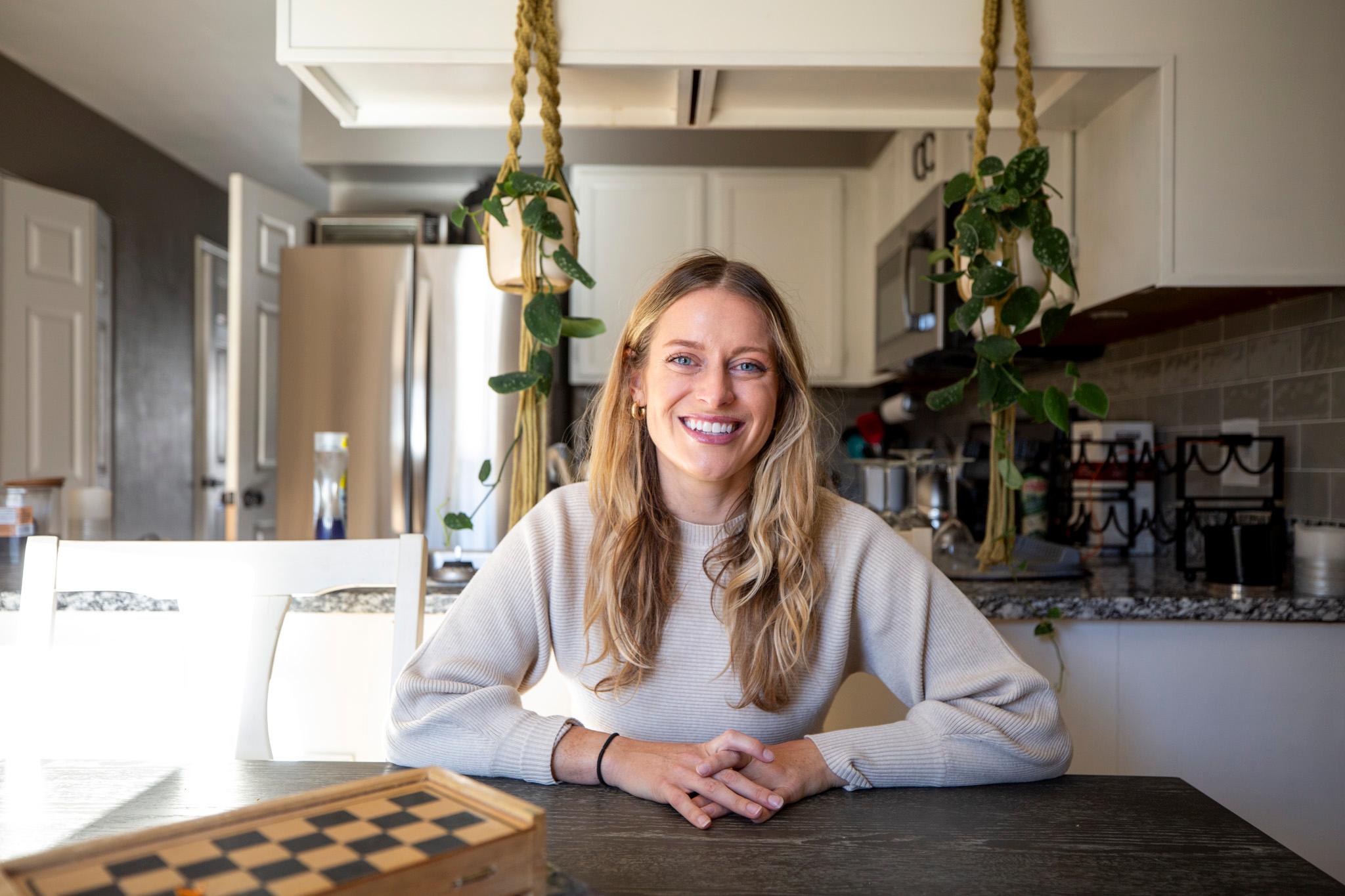 A blonde woman sits at a dark brown kitchen counter, lit by warm sunlight. She is shot from the waist up, wearing a grey sweater and smiling.