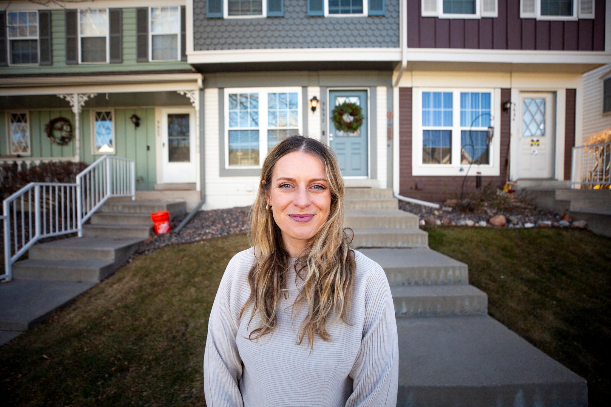 A blonde woman stands in the yard in front of her gray-blue townhome. She is shot from the waist up, wearing a grey sweater and smiling.