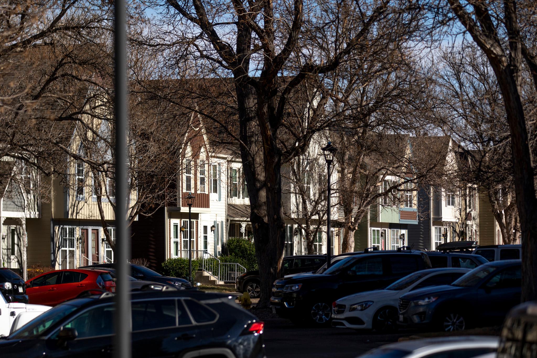 A row of neutral-colored townhomes photographed through a line of trees and parked cars.