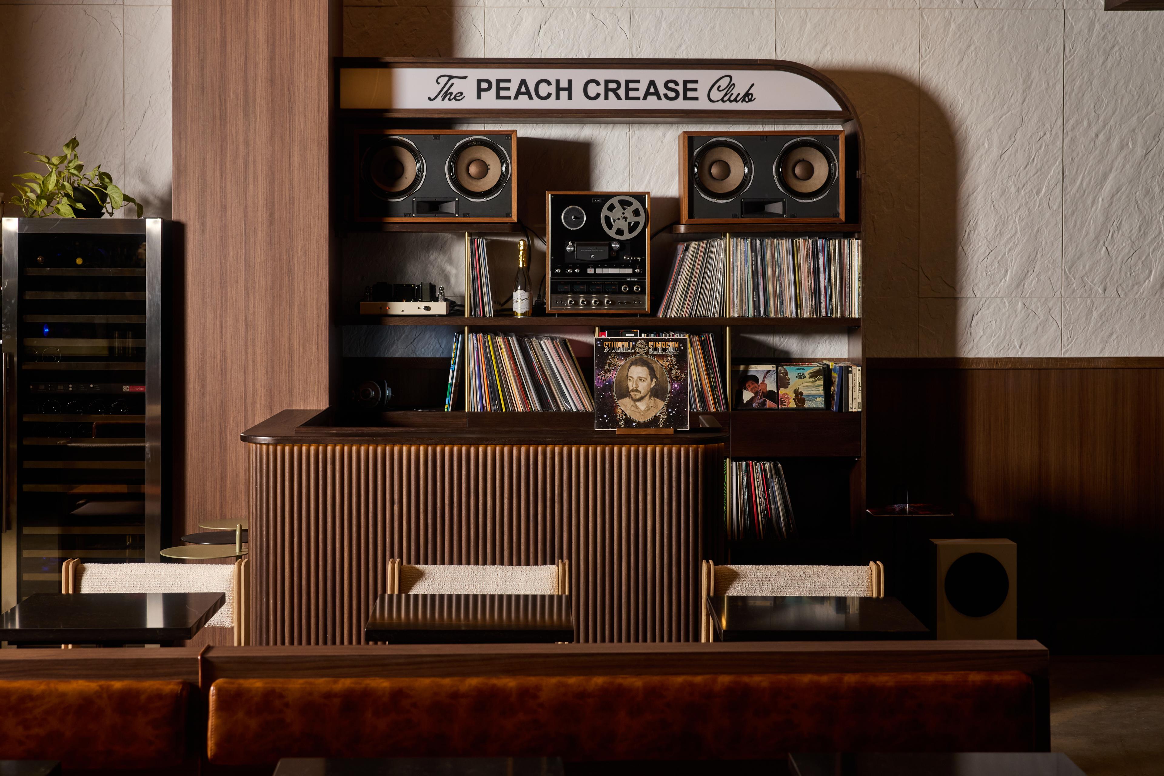 A wooden table with a collection of records and a radio. A white sign atop the shelf reads "The Peach Crease Club."