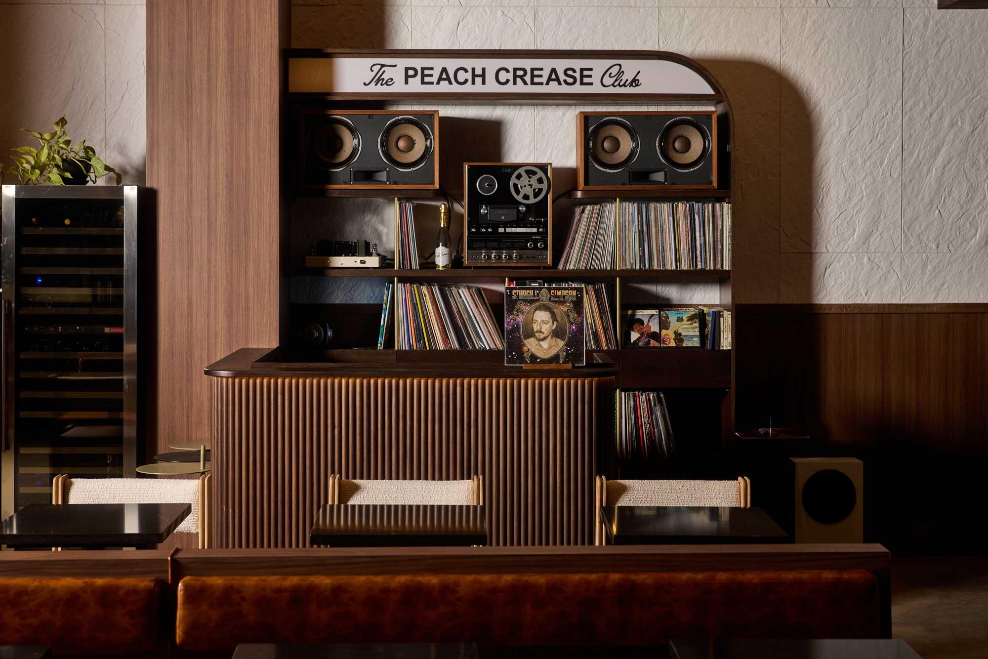 A wooden table with a collection of records and a radio. A white sign atop the shelf reads "The Peach Crease Club."
