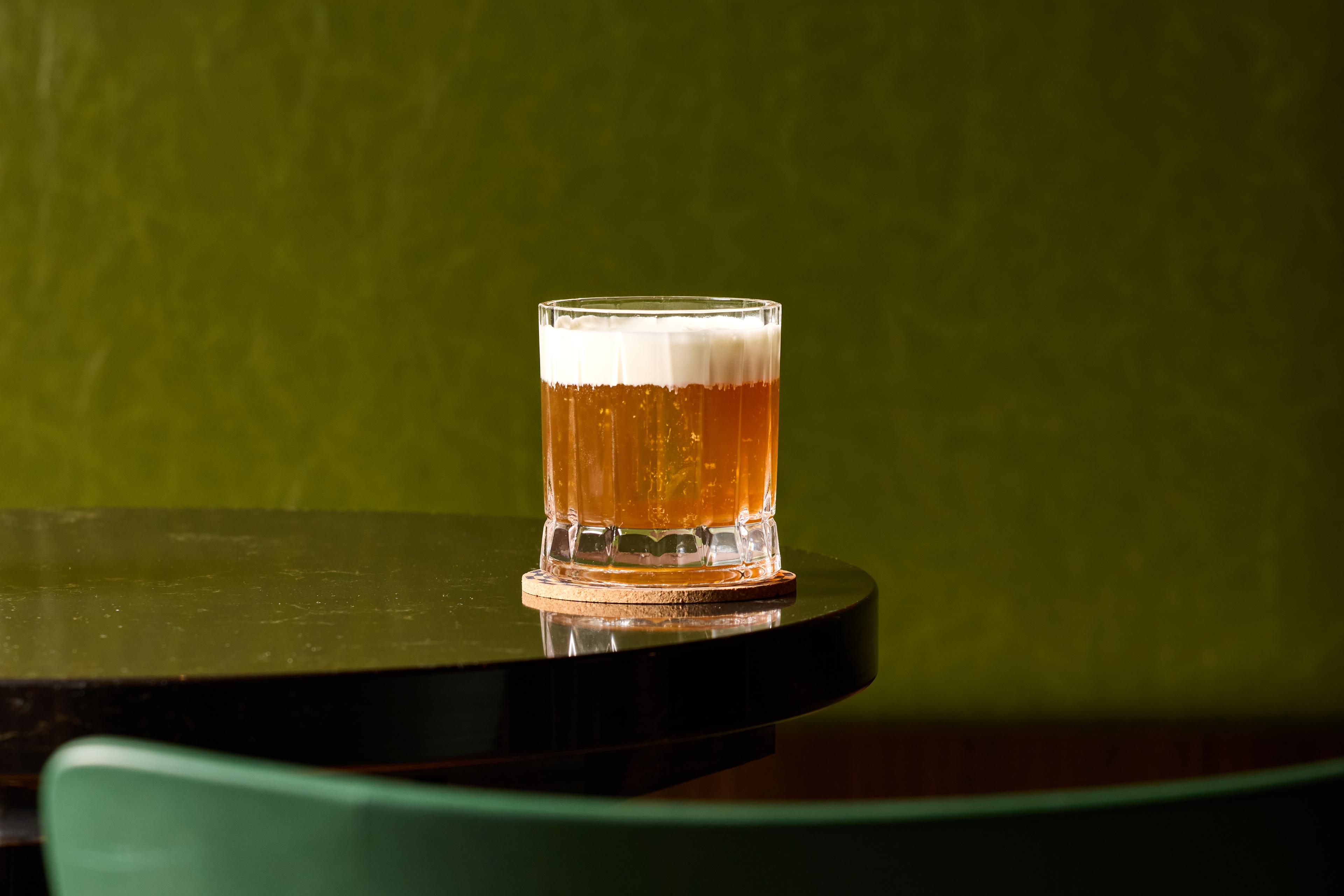 A glass filled with an amber-colored and foamy cocktail rests on a dark wood table.