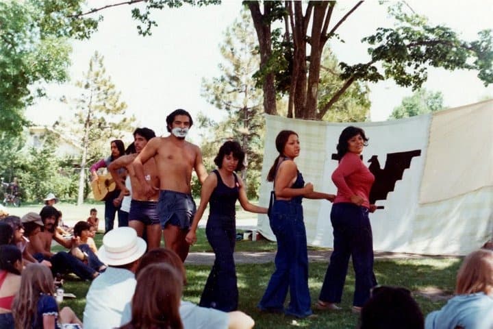 A group of Chicano students perform in front of a black-and-white United Farm Workers flag.