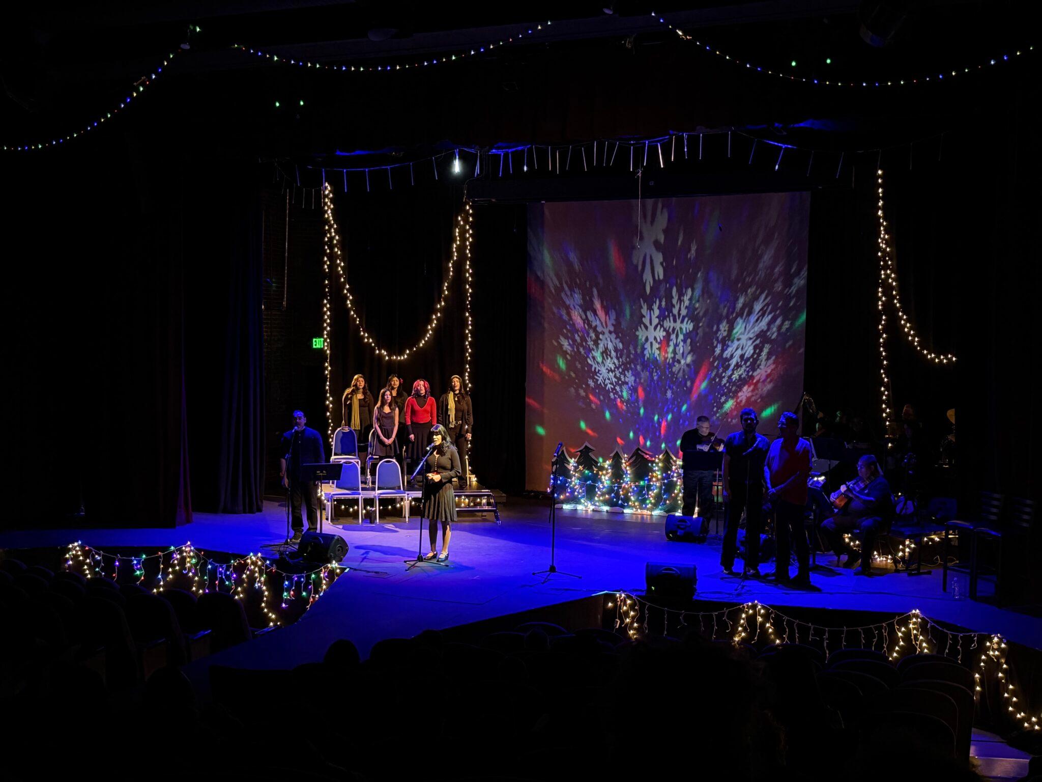 There's a stage, draped in multi-colored Christmas lights with snowflakes and christmas trees projected in the background. Center stage in the spotlight is a young woman at the mic. Also on stage are a chorus of six and about five musicians.