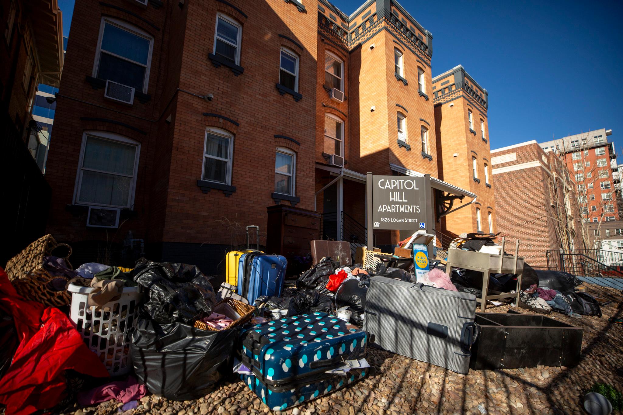 A large pile of items — including a plastic laundry basket, trash bags, suitcases and pieces of furniture — are strewn outside a red brick apartment complex called "Capitol Hill Apartments."