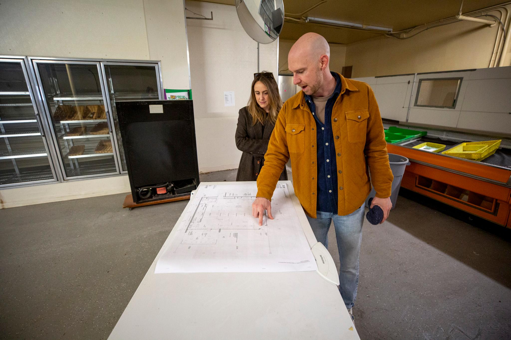 A bald man in an orange jacket and a woman in a brown coat stand in a sparse room with a concrete floor, gazing down at a blueprint on a table.