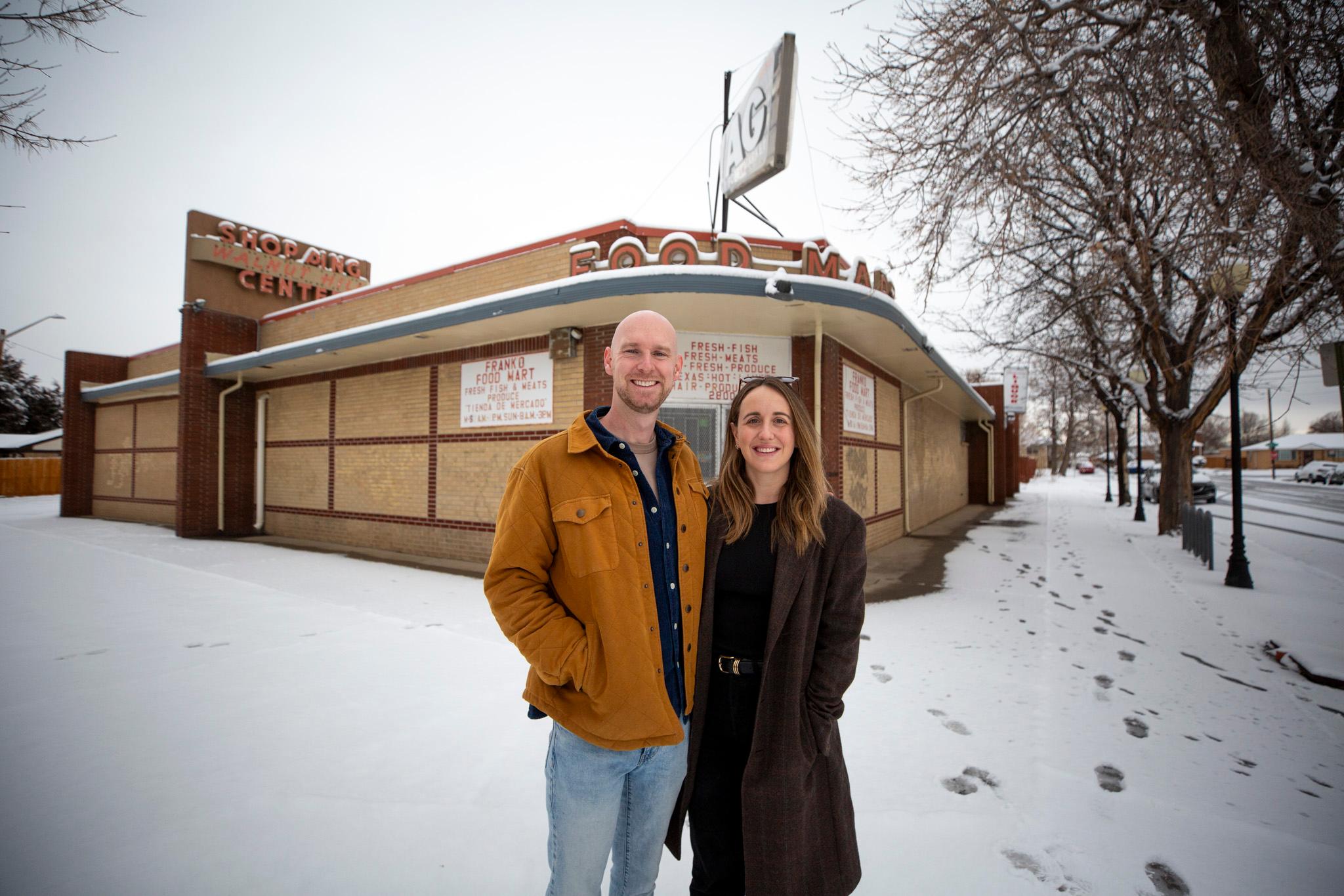 A bald man in an orange jacket and a woman in a brown coat stand in front of a short brown building adorned with an awning and old-timey text that reads "FOOD MART" above a front door.