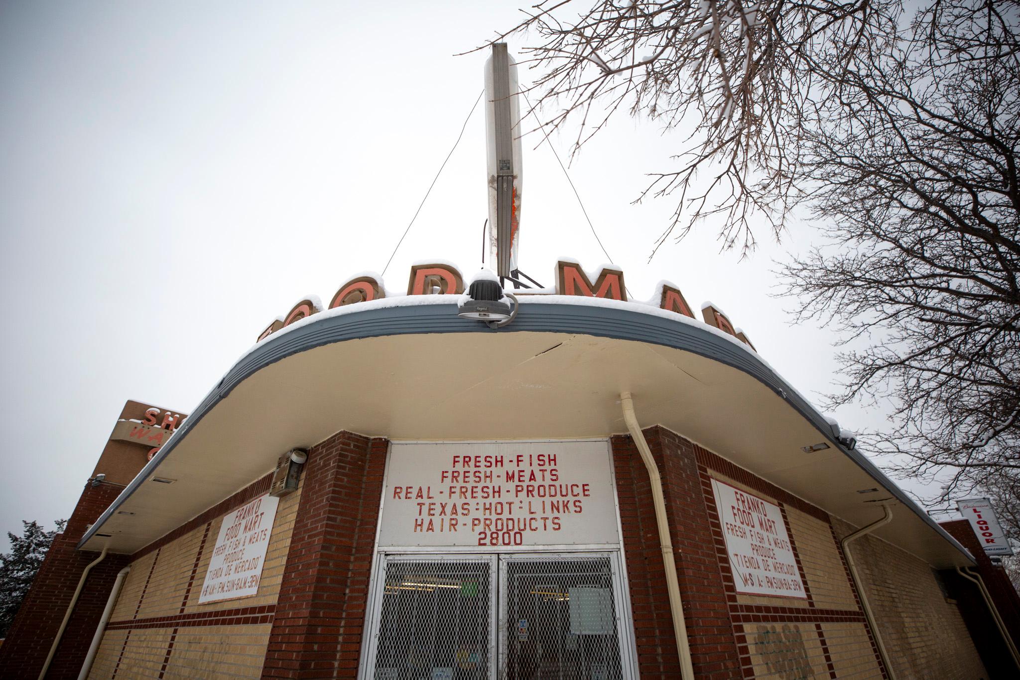 A look up at the front door of short, brown brick building adorned with an awning and old-timey text that reads "FOOD MART." A sign above the door reads "fresh fish, fresh meats, real fresh produce, Texas hot links, hair products."