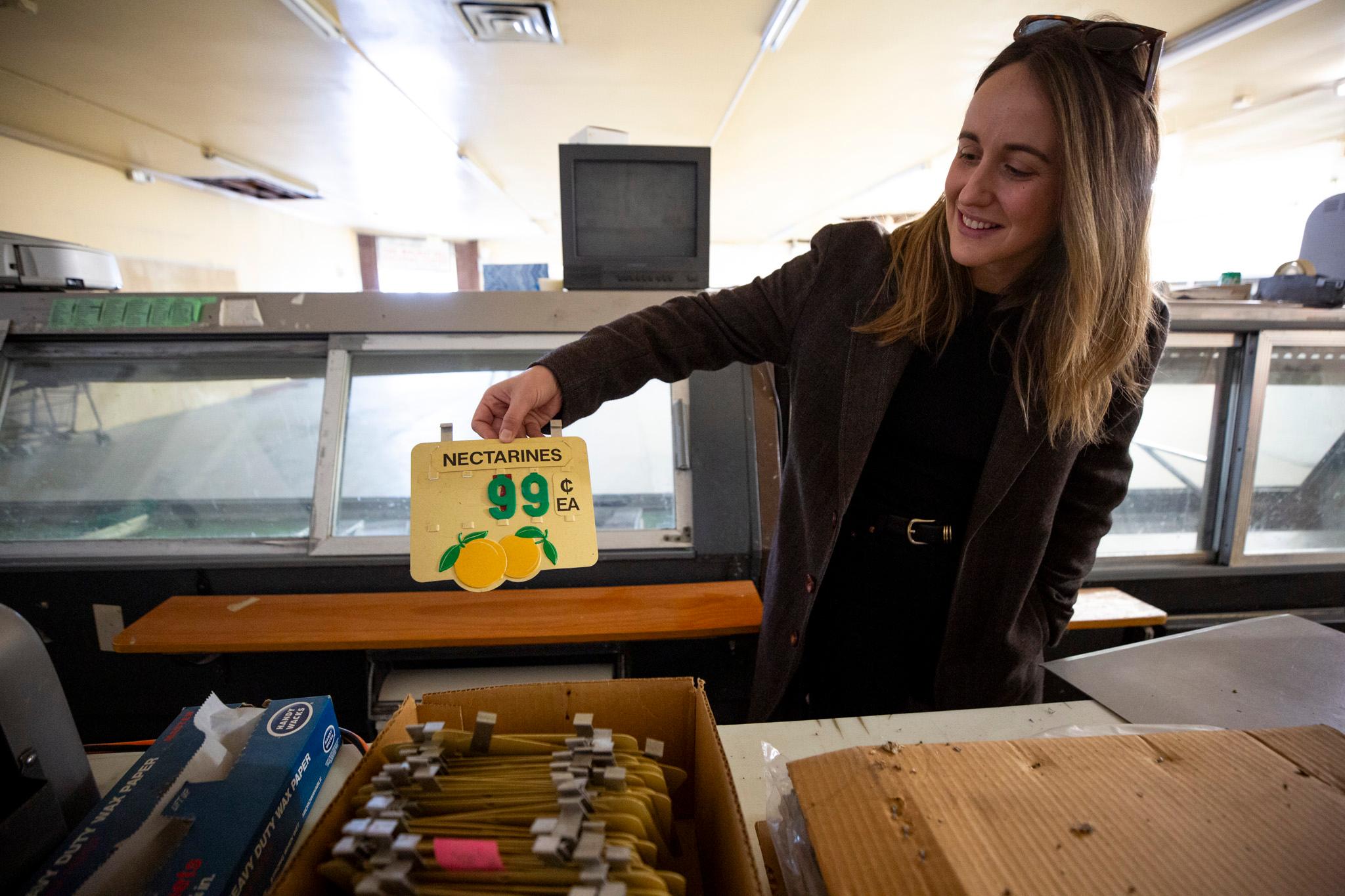 A woman in a brown coat pulls a little white plastic sign out of a box. It reads "nectarines" with big green numbers that show they were "99" cents each.
