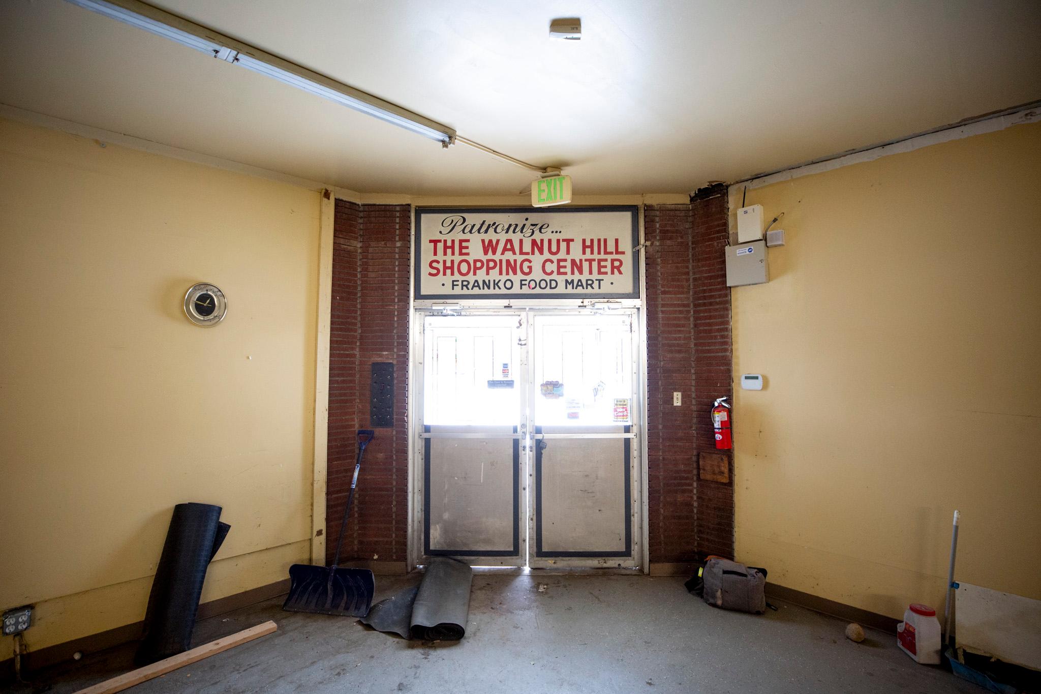 Double doors seen from inside of a warmly painted building, with bright white sunshine glowing in its windows. A sign in old-timey font above reads "Patronize the Walnut Hill Shipping Center, Franko Food Mart."