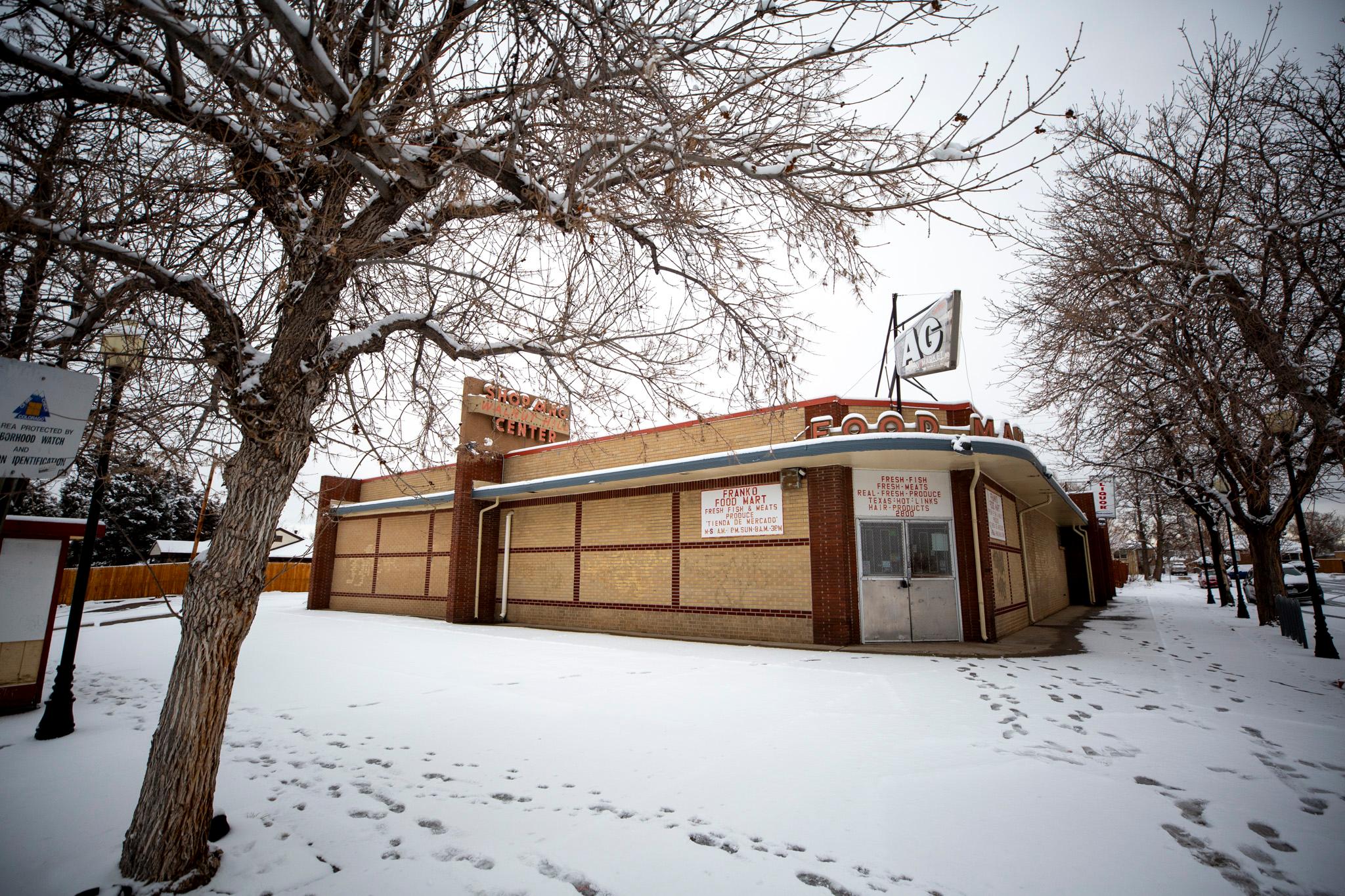 A brown brick building sits on a street corner, which is frosted with a layer of fresh snow. It's lined with an awning and a retro-fonted sign that reads "FOOD MART" over its front door. Barren trees reach into the sky above.
