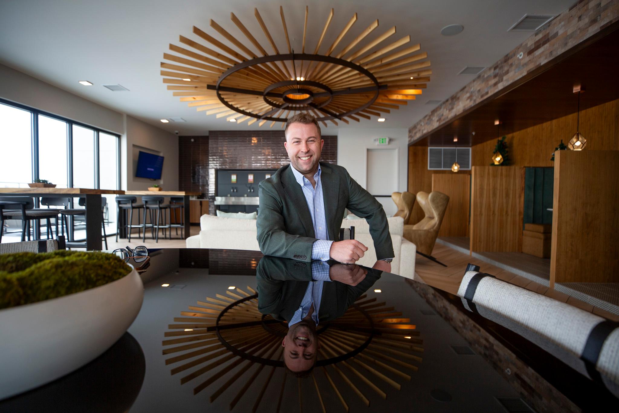 A man in a grey suit jacket smiles at the camera as he sits at a shiny onyx table. A ceiling fixture behind him radiates golden planks like a sunbeam above.