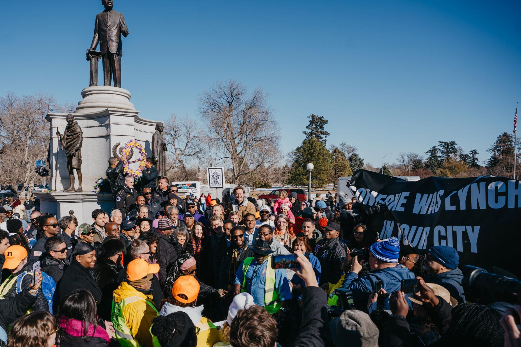 A large crowd stands around a towering statue of Martin Luther King Jr. A banner held up behind them reads "someone was lynched in your city."