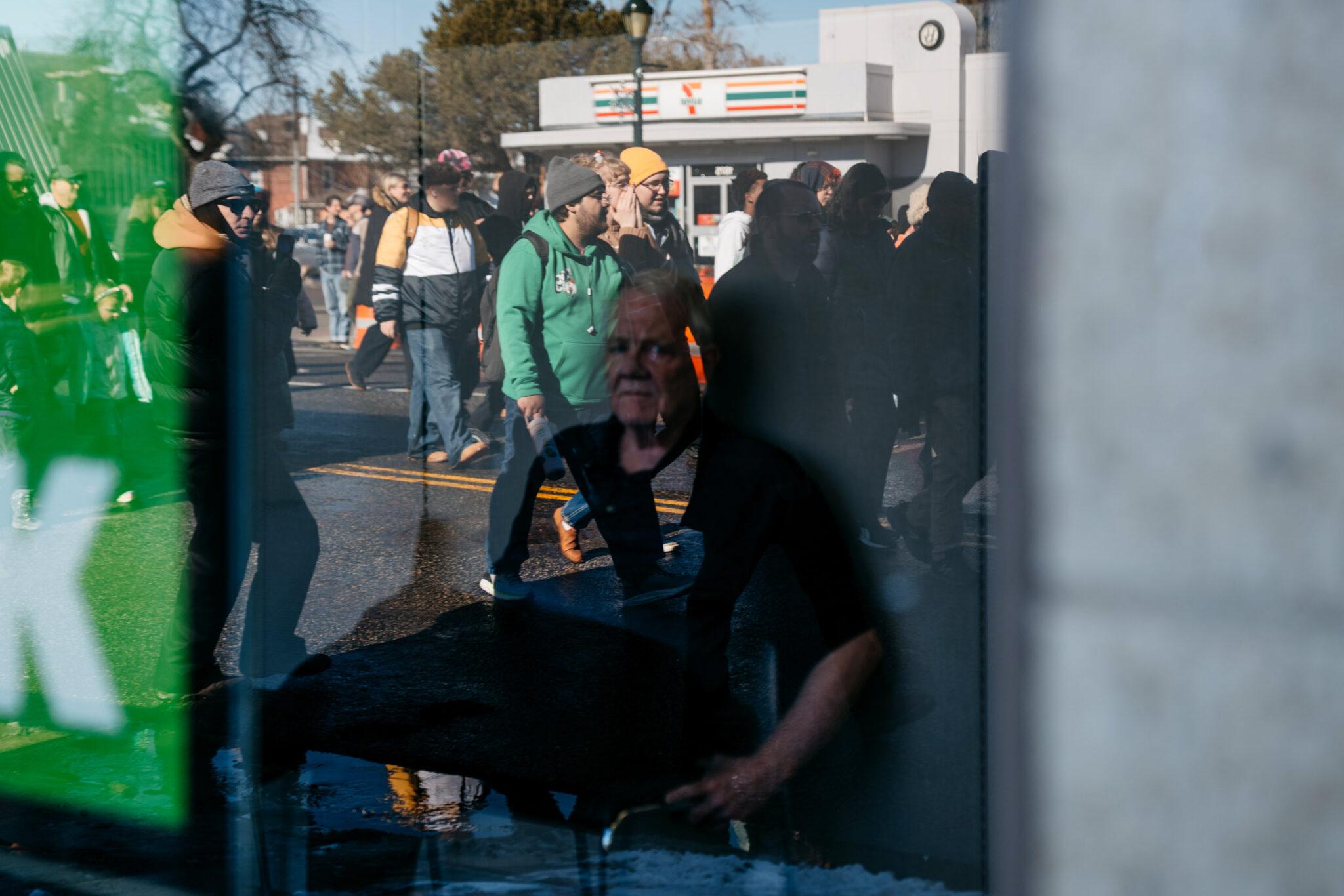 A crowd walking down a street can be seen in the reflection of a glass window, through which we can see an mustached man staring out at them.