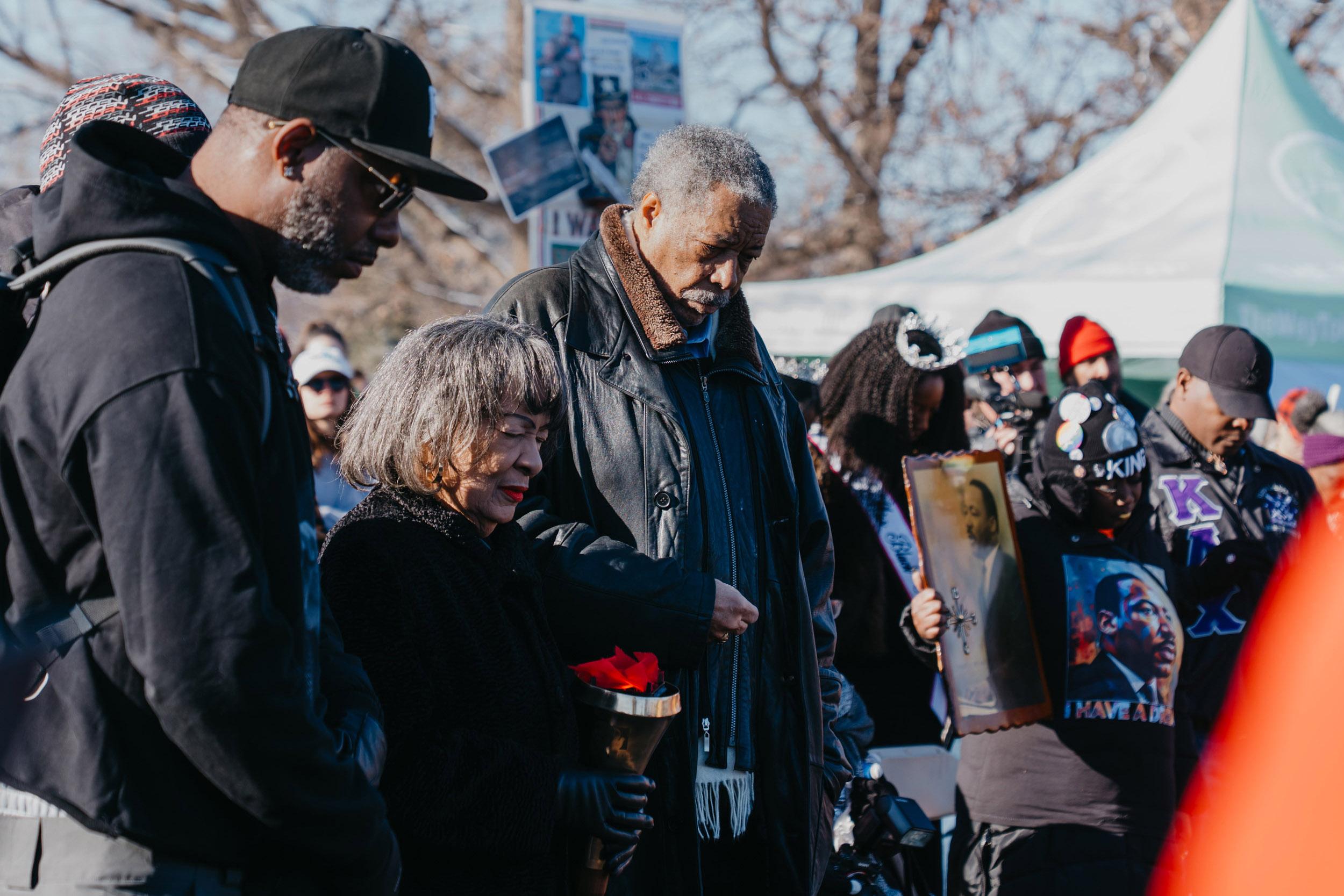 An older couple bow their heads and hold hands in a crowd of people doing the same.
